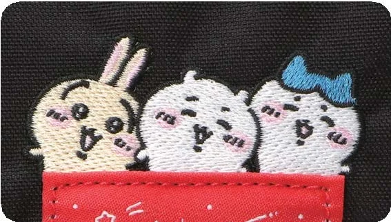 A close-up detail shot of the Manhattan Portage x Chiikawa collaboration embroidery featuring three beloved Chiikawa characters peeking out from behind the brand’s signature red woven label. Set against textured black canvas fabric, the stitched artwork highlights soft pastel tones, expressive blush cheeks, and playful facial expressions, blending kawaii character culture with Manhattan Portage’s rugged utilitarian bag construction