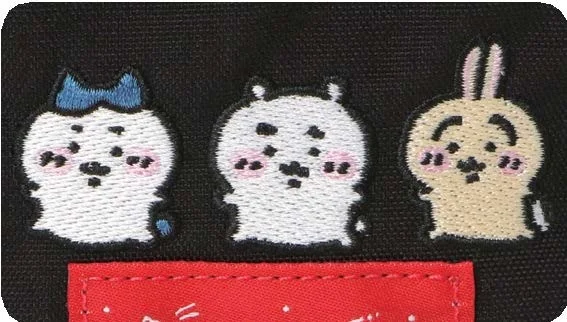 A macro-style embroidered detail from the Manhattan Portage x Chiikawa collection showing three Chiikawa characters stitched above the label placement on black canvas material. The design features soft cream and white threadwork, rosy cheek accents, and a small blue bow detail, creating a play contrast against the rugged fabric texture and Manhattan Portage’s iconic bright red tag below