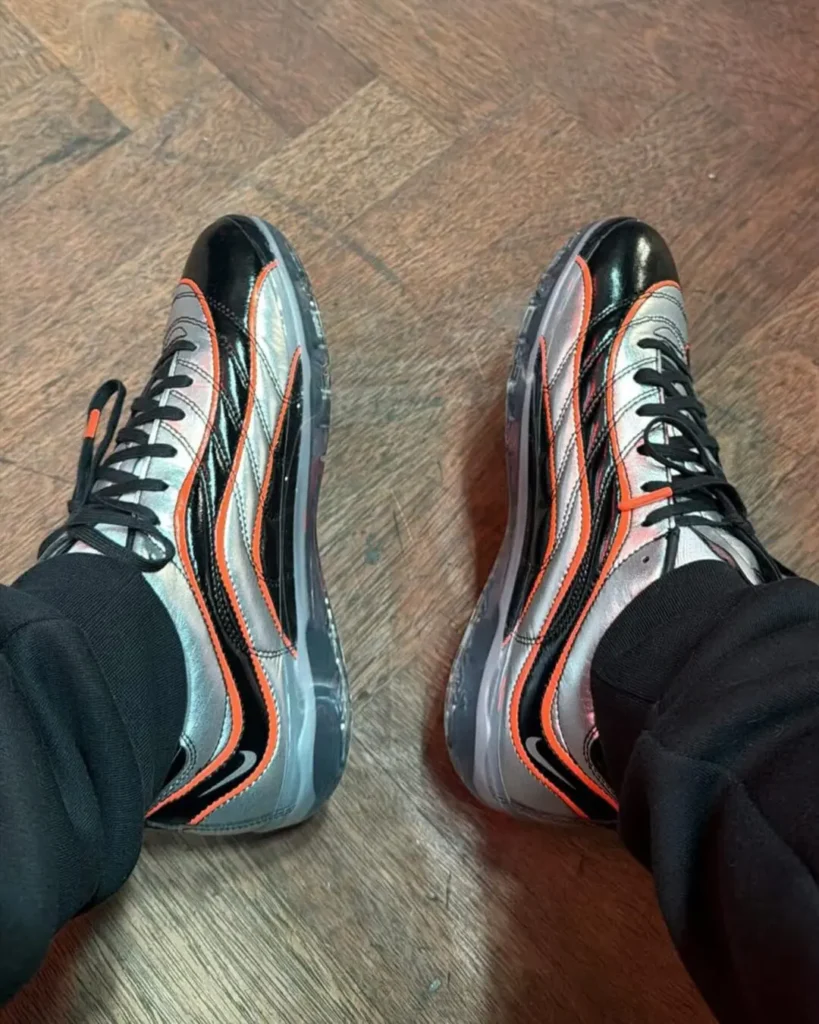 Top-down on-foot view of a pair of metallic silver Nike shoe featuring glossy black wave-shaped overlays and bright orange piping, finished with black laces and translucent soles, worn with black pants on a dark wood floor