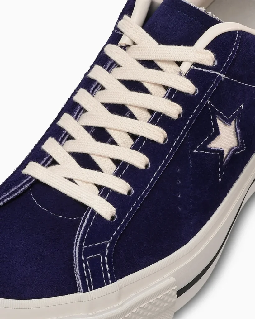 Macro detail shot of the Converse TimeLine ONE STAR J VTG “Purple,” highlighting the rich textured purple suede upper, vintage off-white cotton laces, and contrast white stitching surrounding the signature cut-out One Star logo. The close-up emphasizes the premium Made-in-Japan construction and archival material finish, paired with a clean white vulcanized foxing and classic black stripe detail