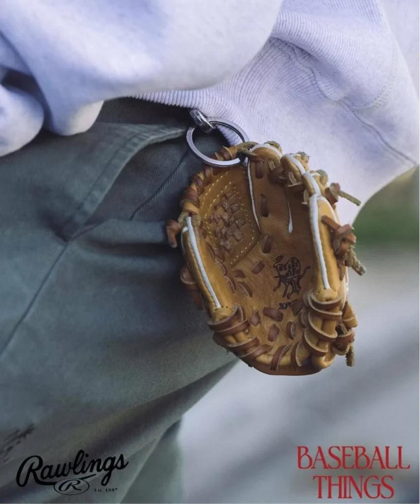 Lifestyle editorial image of a miniature Rawlings baseball glove keychain crafted from tan leather with woven detailing and traditional lacing, clipped onto olive trousers beneath a grey sweatshirt as part of the “Baseball Things” accessory campaign