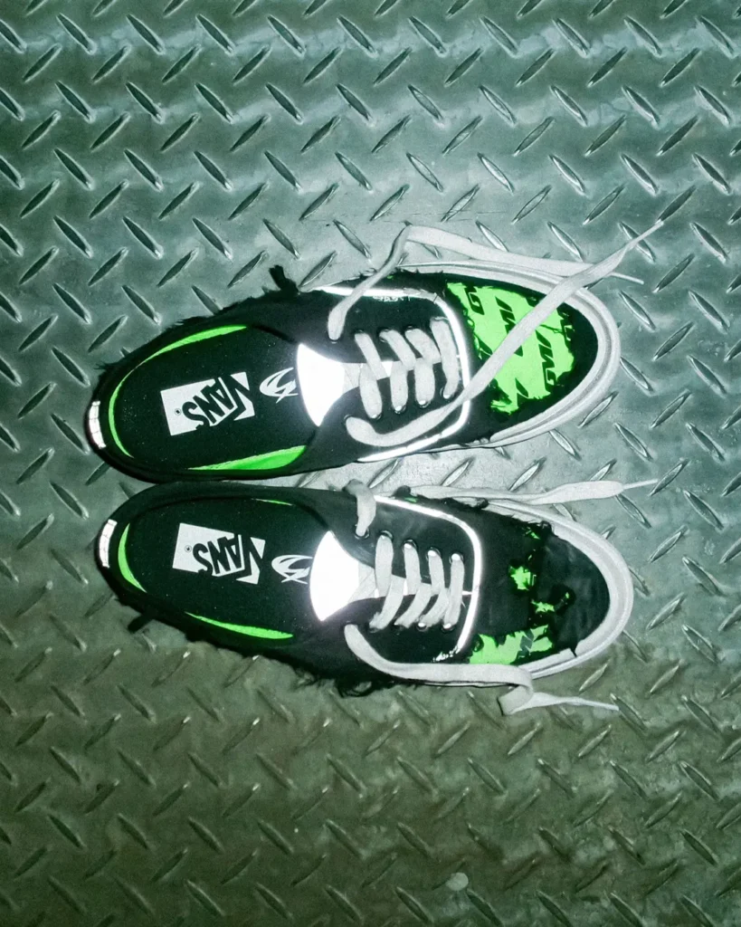 Overhead view of the Vans x GUCCIMAZE PREMIUM AUTHENTIC shoe showcasing the collection's distressed black upper, reflective detailing, and exposed neon green graphics against a textured metal floor