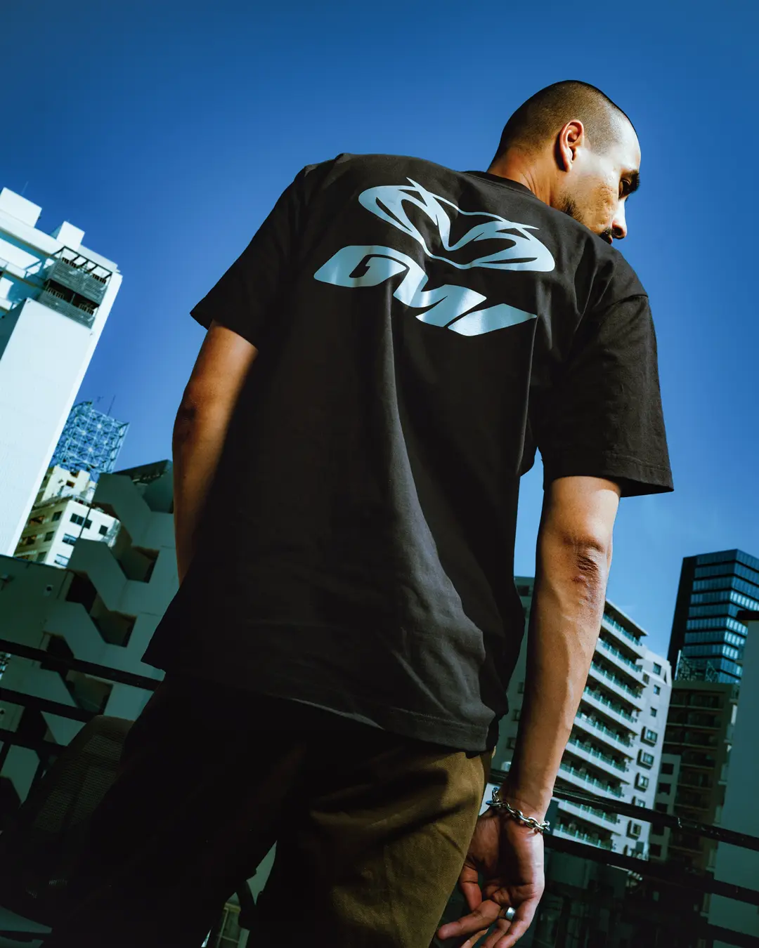 Model wearing the Vans x GUCCIMAZE collision apparel, highlighting the oversized reflective graphic logo printed across the back of a black T-shirt against an urban