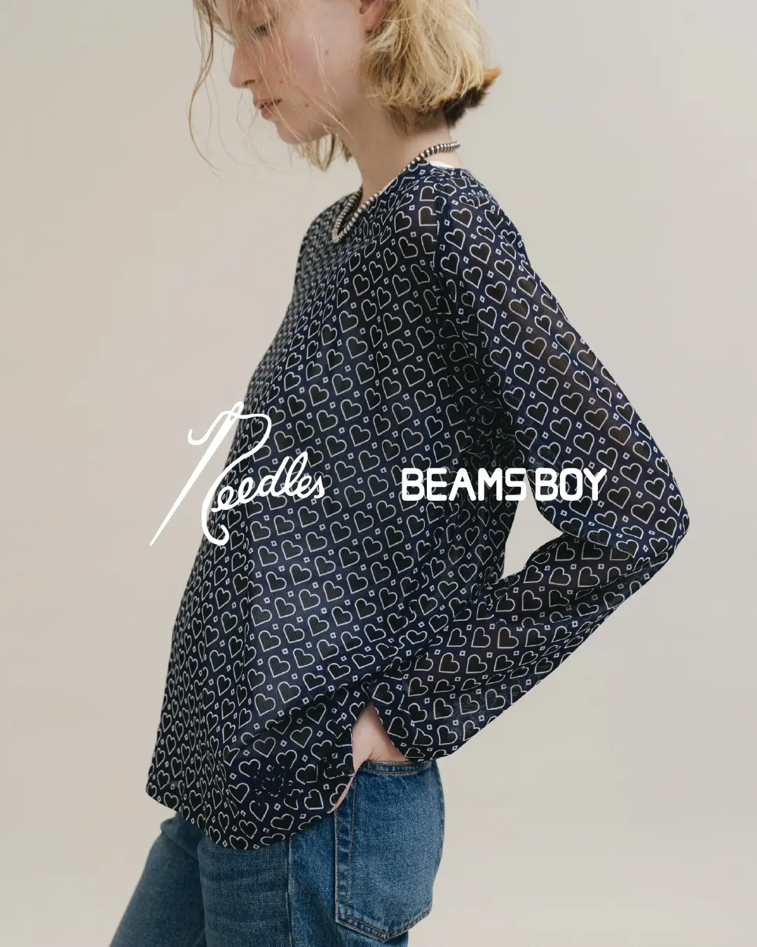 Model wearing a navy long-sleeve Needles x BEAMS BOY top covered in an all-over heart motif, styled with blue denim jeans and silver jewelry in a minimalist studio set