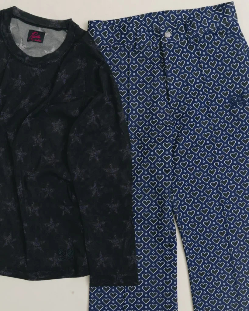 Flat-lay of the Needles x BEAMS BOY collection featuring a black long-sleeve top with tonal butterfly graphics alongside blue heart-patterned trousers