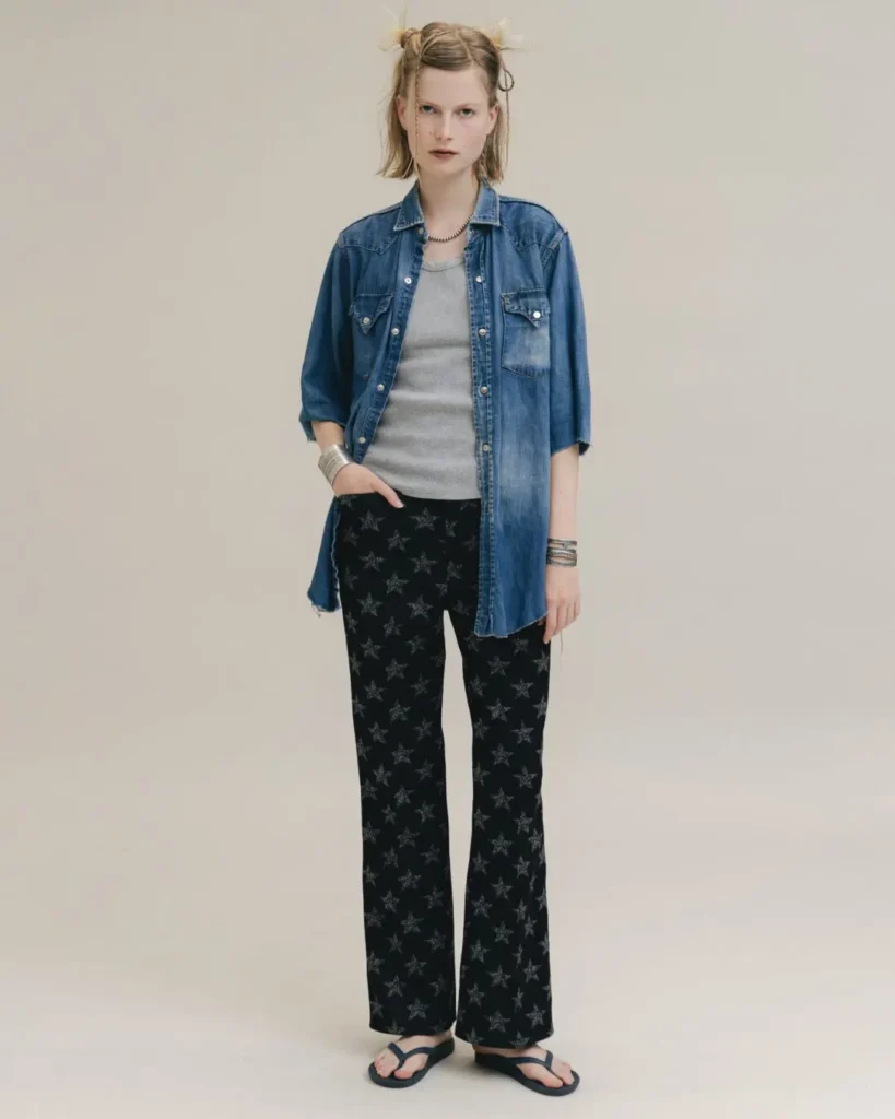 Full-body look from the Needles x BEAMS BOY collaboration featuring a cropped denim shirt, star-patterned black trousers, silver accessories, and flip-flops against a clean studio backdrop