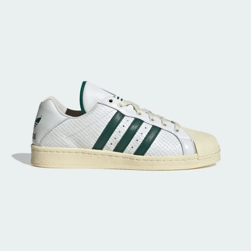 Side profile of the adidas Originals ULTRASTAR Made in Germany “White/Green” sneaker featuring textured white premium leather, dark green Three Stripes, an aged cream shell toe and midsole, tonal laces, and heritage-inspired low-top construction against a light grey background
