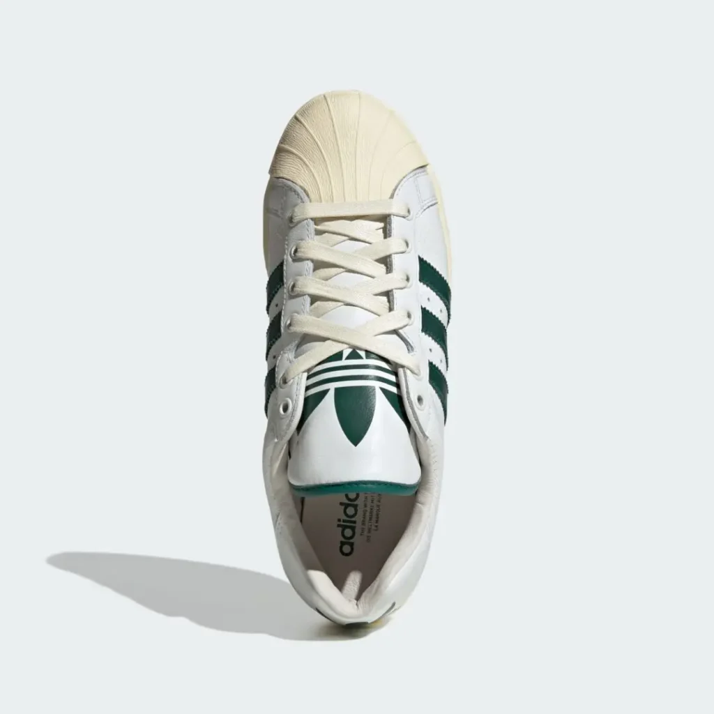 Top-down view of the adidas Originals ULTRASTAR Made in Germany “White/Green” showcasing cream flat laces, an oversized green Trefoil tongue graphic, premium white leather construction, signature shell toe detailing, and deep green Three Stripes against a clean light grey backdrop