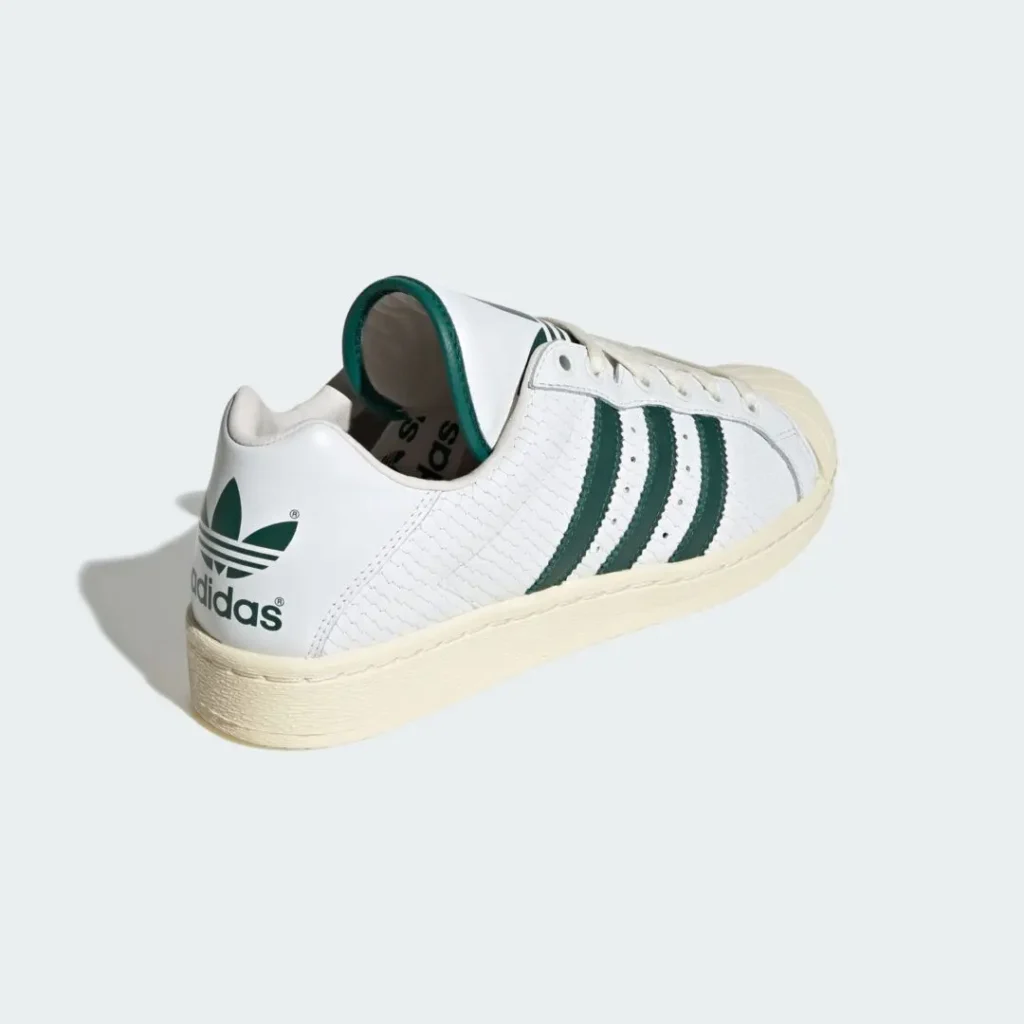 Rear three-quarter view of the adidas Originals ULTRASTAR Made in Germany “White/Green” highlighting the oversized green Trefoil heel branding, textured white leather upper, dark green Three Stripes, vintage cream midsole, and classic shell-toe silhouette with elevated heritage detailing