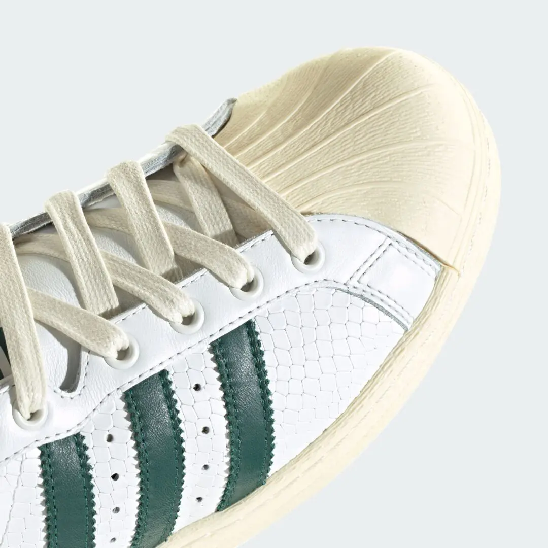 Macro close-up of the adidas Originals ULTRASTAR Made in Germany “White/Green” highlighting the vintage cream shell toe, textured white leather upper, deep green serrated Three Stripes, cream flat laces, and aged midsole finish that emphasizes the shoe premium heritage construction