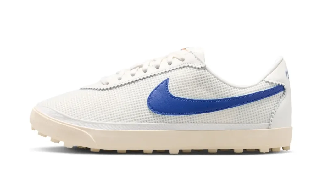 Side-profile view of a white mesh Nike low-top sneaker featuring a bold royal blue leather Swoosh, cream-toned midsole, and rugged gum waffle outsole, balancing retro running heritage with minimalist contemporary style