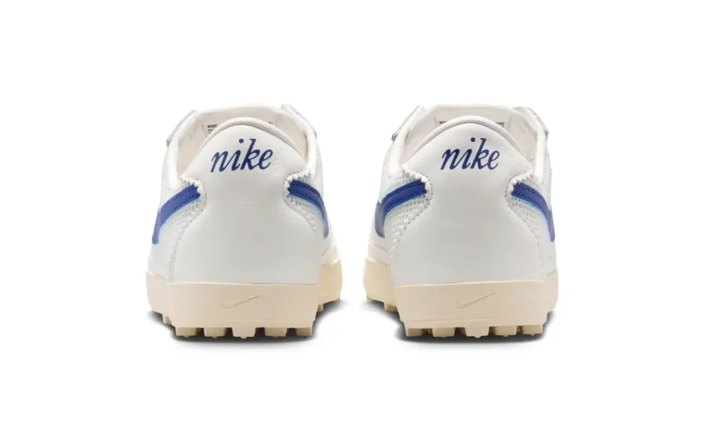 Rear view of the white Nike low-profile shoe featuring blue embroidered heel branding, extended royal blue Swoosh tails, cream-toned midsoles, and exposed waffle traction detailing inspired by vintage track footwear