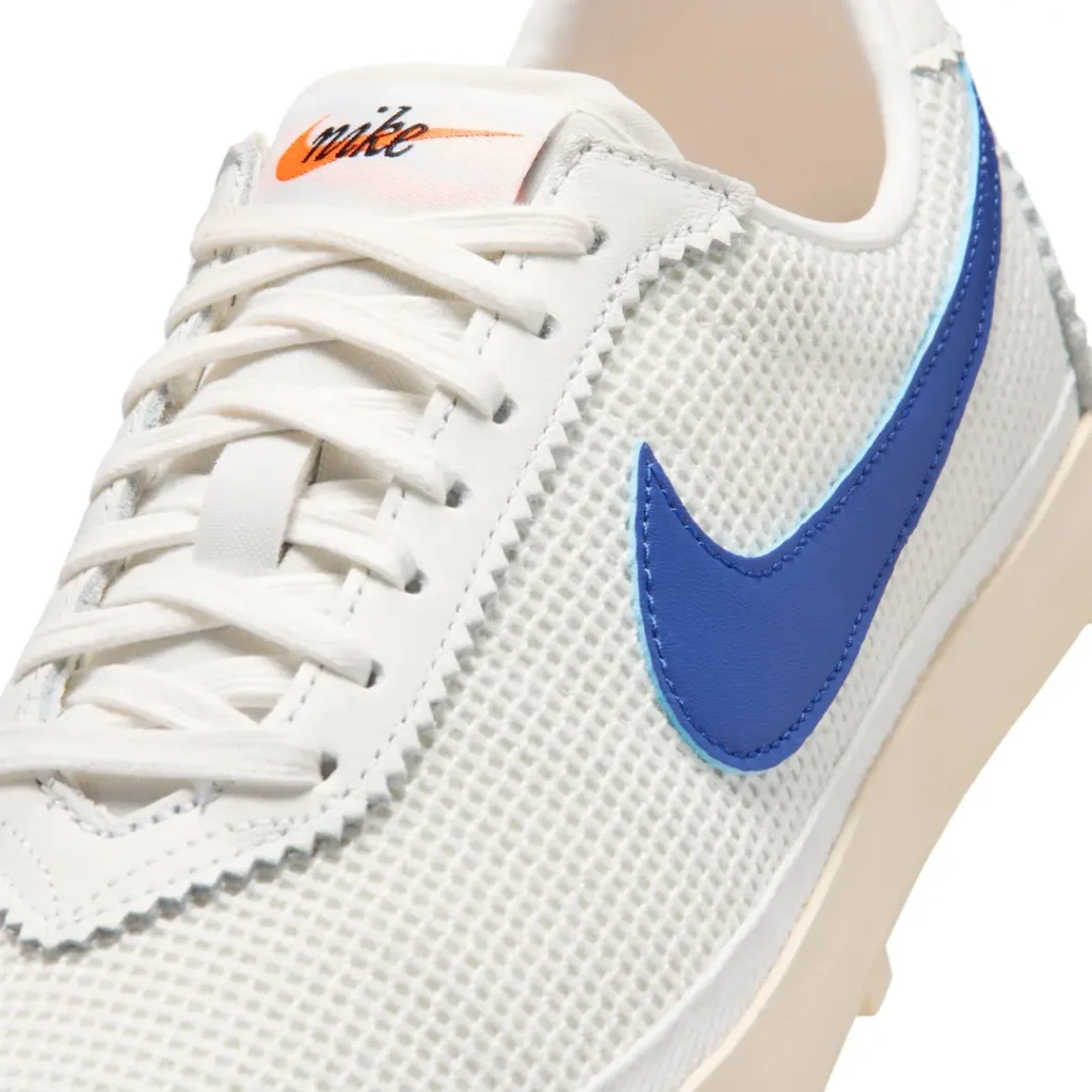 Close-up heel detail of the Nike shoe featuring textured white mesh construction, a royal blue leather Swoosh tail with icy blue edging, cream vintage-style midsole, and chunky waffle traction lugs underneath