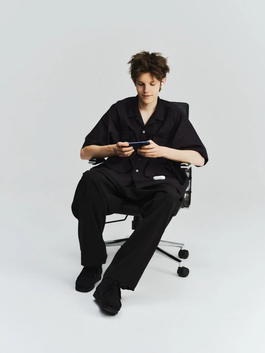 Minimal studio fashion portrait of a model seated in a black office chair wearing an oversized black short-sleeve utility shirt with matching relaxed trousers and black shoes. Looking down at a handheld device, the styling emphasizes clean lines, oversized proportions, and understated contemporary workwear against a light gray backdrop