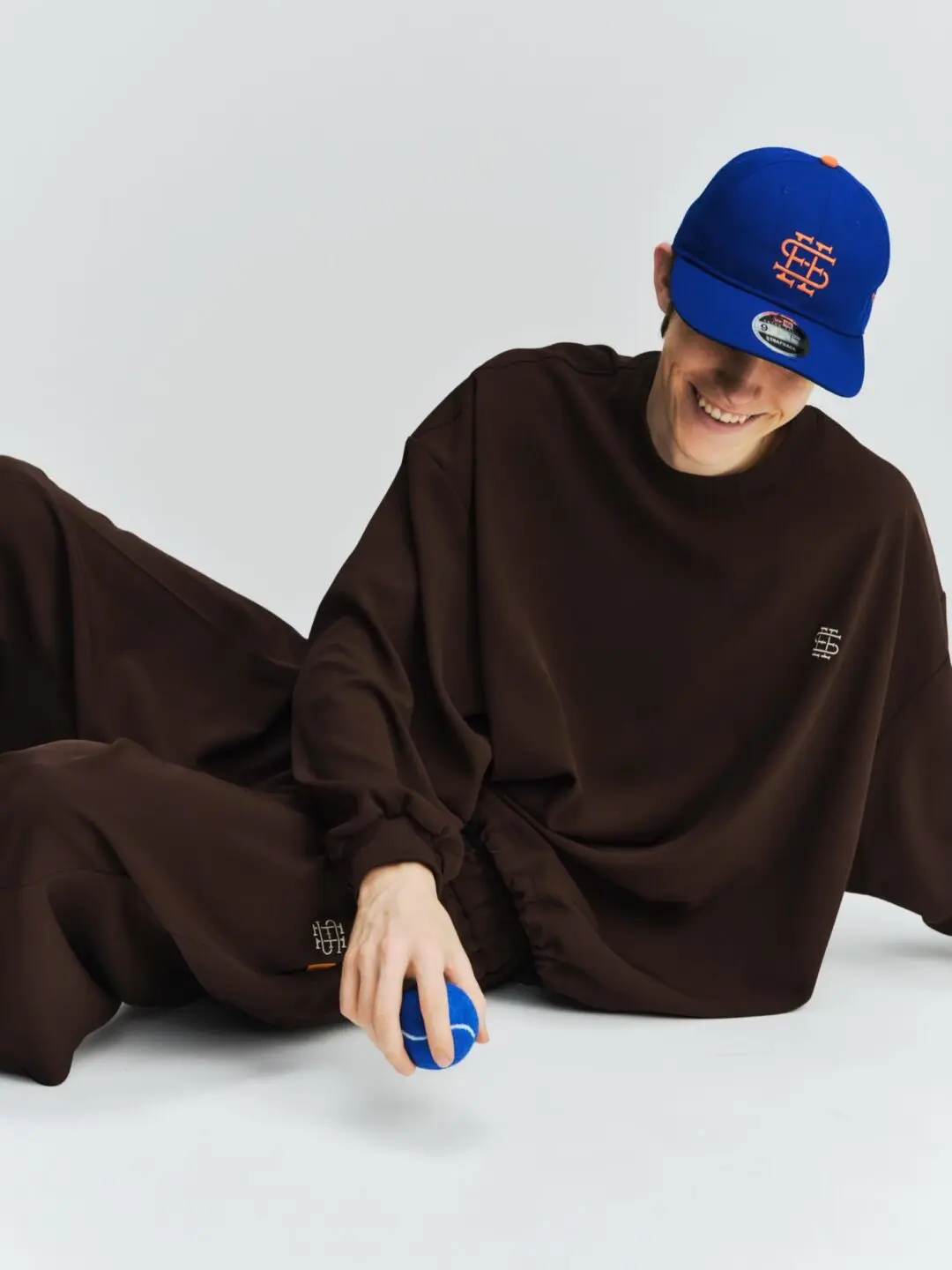 Studio fashion portrait of a model reclining on a light gray floor wearing an oversized dark brown SEE SEE sweatshirt and matching relaxed pants. The look is paired with a vivid royal blue cap featuring an orange embroidered monogram, while the model casually holds a matching blue ball and smiles downward, creating a playful contrast against the minimal backdrop