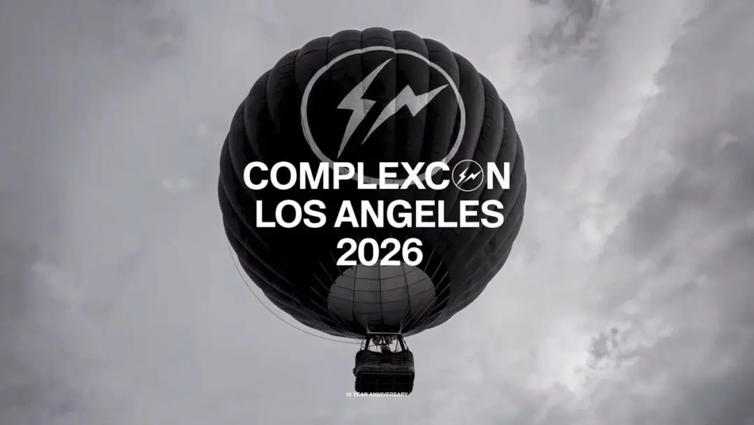 Low-angle promotional image of a single oversized matte-black hot air balloon floating against a cloudy grey sky, carrying a merged Fragment lightning-bolt insignia and event branding. Bold white typography across the center reads “COMPLEXCON LOS ANGELES 2026,” while a subtle “10 YEAR ANNIVERSARY” mark appears near the basket, reinforcing the campaign’s milestone status. The stark monochrome palette and elevated perspective create a cinematic atmosphere that feels equal parts luxury campaign, streetwear teaser, and culture event announcement
