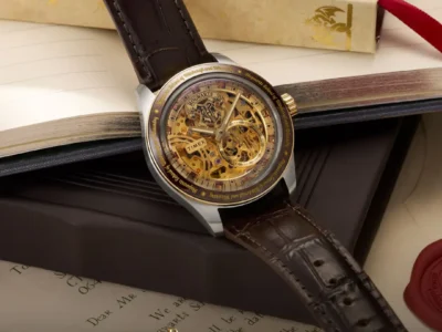 Close-up campaign image of the Timex x Harry Potter and the Philosopher's Stone Waterbury Skeleton Automatic watch featuring a gold-toned exposed mechanical dial, Hogwarts crest detailing, brown crocodile-grain leather strap, and engraved bezel resting atop an open wizard-themed book beside parchment-style packaging