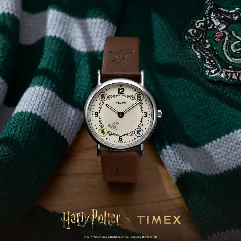 Campaign image of the Timex x Harry Potter and the Philosopher's Stone Weekender watch featuring a cream dial with Hogwarts house-inspired detailing, Sorting Hat graphic at 6 o’clock, black numeral markers, polished silver-tone case, and brown leather strap embossed with Sorting Hat motifs, styled against a green-and-silver Slytherin scarf backdrop
