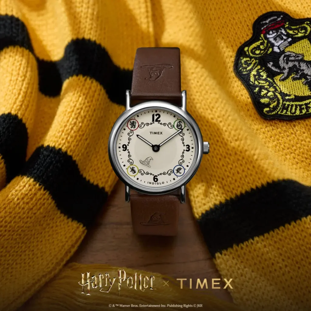 Campaign image of the Timex x Harry Potter and the Philosopher's Stone Weekender watch featuring a cream dial with Hogwarts house-inspired detailing, Sorting Hat graphic at 6 o’clock, black numeral markers, polished silver-tone case, and brown leather strap embossed with Sorting Hat motifs, styled against a yellow-and-black Hufflepuff scarf backdrop
