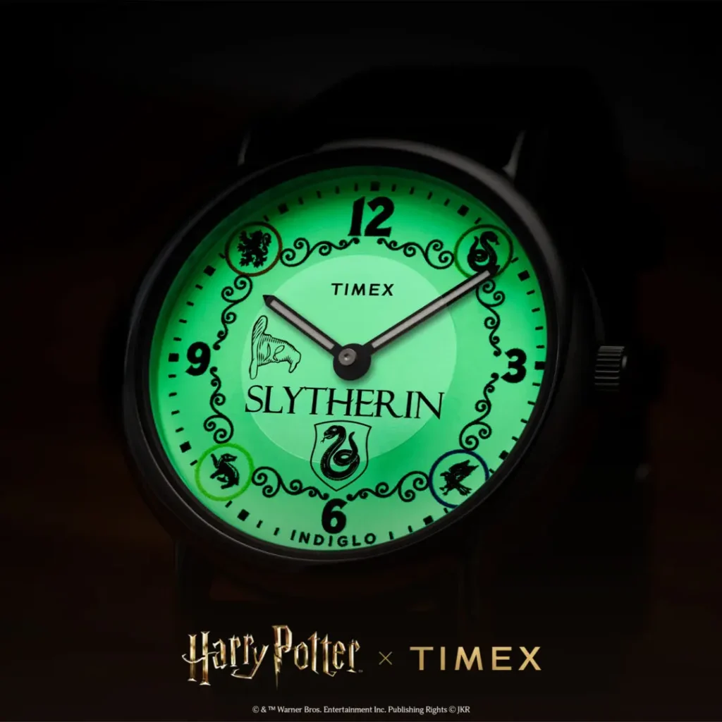 Close-up low-light campaign image of the Timex x Harry Potter and the Philosopher's Stone Slytherin Weekender watch illuminated with green INDIGLO® backlighting, featuring Slytherin crest branding, Sorting Hat graphic, ornate black detailing, and bold numeral markers against a dark background