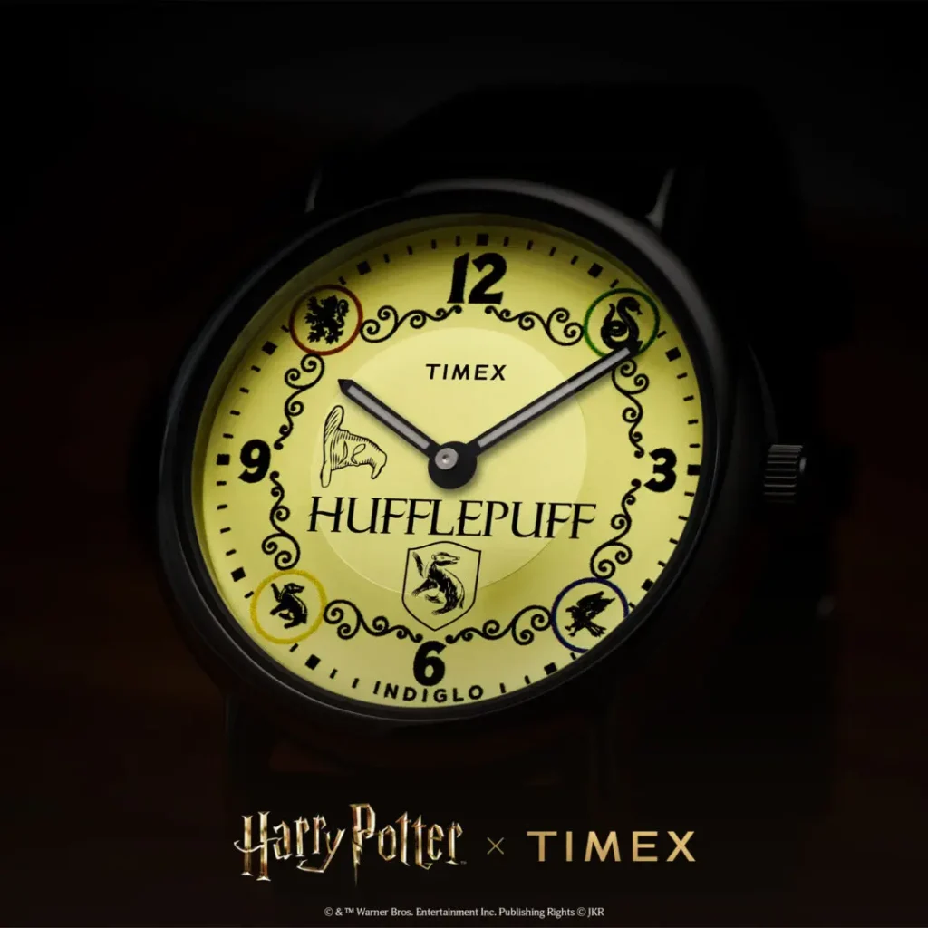 Close-up low-light campaign image of the Timex x Harry Potter and the Philosopher's Stone Hufflepuff Weekender watch illuminated with yellow INDIGLO® backlighting, featuring Hufflepuff crest branding, Sorting Hat graphic, ornate black detailing, and bold numeral markers against a dark background