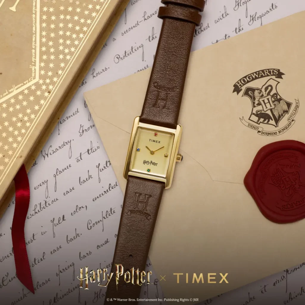 Lifestyle campaign image of the Timex x Harry Potter and the Philosopher's Stone Hailey Hogwarts watch featuring a rectangular gold-tone case, brushed champagne dial with house-colored crystal markers, and embossed brown leather strap, styled atop handwritten parchment, Hogwarts crest stationery, and wax-sealed wizarding correspondence