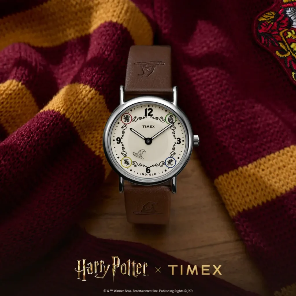 Campaign image of the Timex x Harry Potter and the Philosopher's Stone Weekender watch featuring a cream dial with Hogwarts house-inspired detailing, Sorting Hat graphic at 6 o’clock, black numeral markers, polished silver-tone case, and brown leather strap embossed with Sorting Hat motifs, styled against a Gryffindor scarf backdrop
