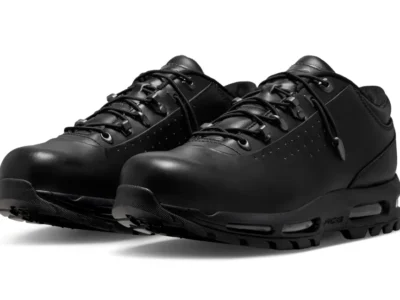 Pair of all-black low-cut trail-inspired shoes photographed against a light gray background. The design features a smooth matte upper with minimal perforation detailing, rounded toe shape, speed-style lacing hardware with loop eyelets, padded collars, and a rugged outsole. Visible cushioning windows and subtle technical branding along the midsole give the silhouette an understated utility and outdoorshoe aesthetic