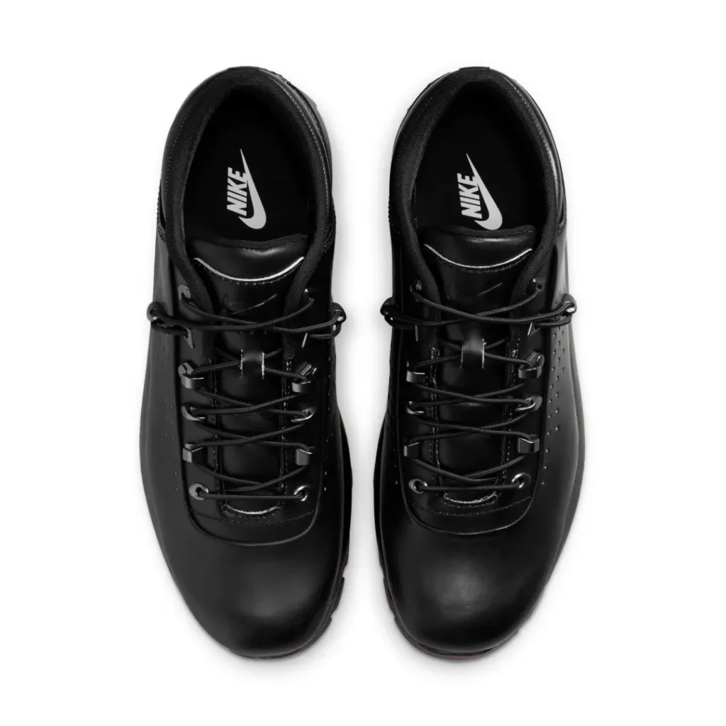 Top-down view of a pair of all-black low-top technical shoes displayed on a light gray background. The design highlights smooth rounded toe boxes, minimalist perforation along the sides, and rugged speed-lace hardware with elastic-style lacing. Padded collars and black interiors feature contrasting white logo branding on the insoles, reinforcing the clean monochrome, utility-driven aesthetic