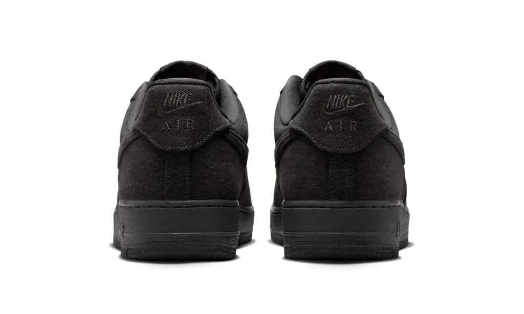 Rear view of the Nike Air Force 1 Low “Denim/Anthracite/Medium Ash,” showcasing tonal anthracite denim construction across the heel panels, embroidered Nike Air branding on the back tabs, padded collars, visible center heel seam detailing, and a matching dark Air sole unit set against a clean light grey studio background