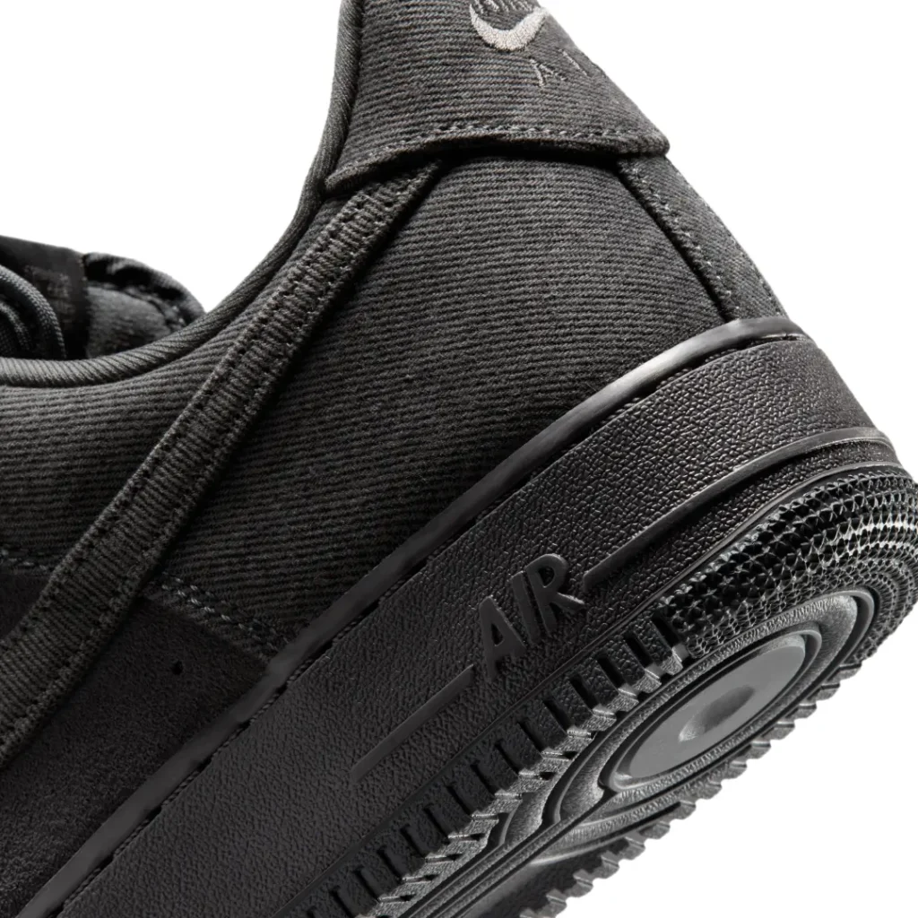 Macro close-up of the Nike Air Force 1 Low “Denim/Anthracite/Medium Ash,” focusing on the heel section with textured anthracite denim construction, tonal embroidered Nike Air branding, black cupsole with embossed AIR lettering, and the classic circular outsole traction pattern rendered in a matching monochrome finish