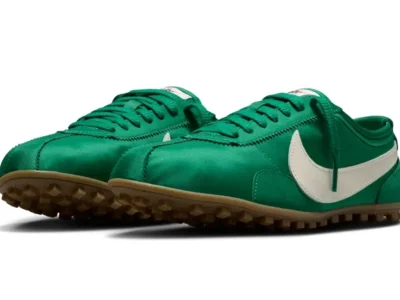 A pair of Nike Cortez-style turf shoe in vivid emerald green satin sits against a light grey backdrop, featuring oversized white leather Swooshes, tonal green laces, and gum rubber outsole traction. The low-profile silhouette blends retro track aesthetics with indoor football-inspired detailing, while the glossy textile upper gives the classic design a more fashion-oriented finish
