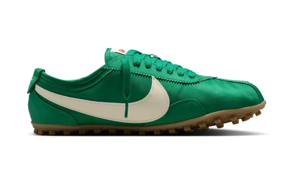 Lateral side view of a satin-finished Nike Cortez-inspired turf shoe in rich emerald green with an oversized sail-white Swoosh and gum rubber studded outsole. The streamlined silhouette combines vintage running shoe proportions with football training functionality, highlighted by tonal laces, zigzag eyestay trim, and a glossy textile upper