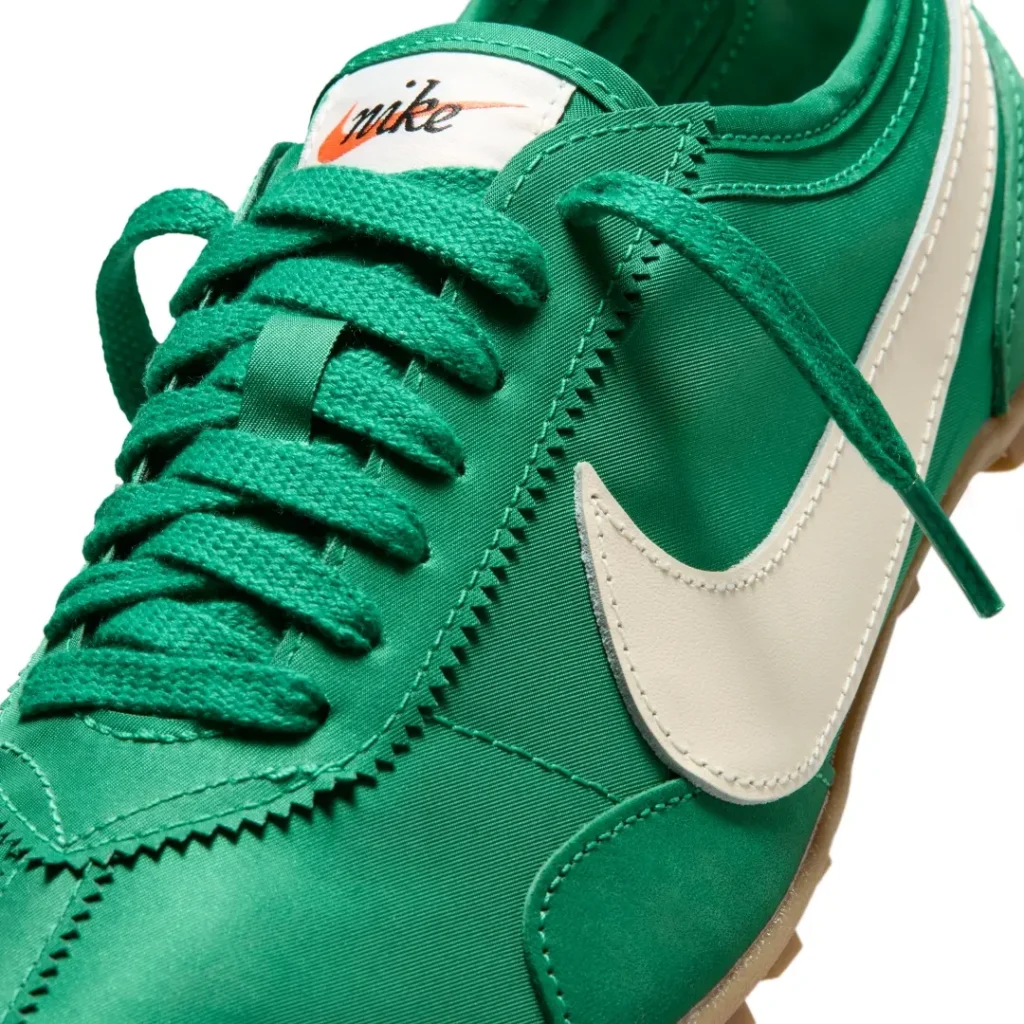 Close-up detail of the emerald green Nike Cortez-inspired turf sneaker highlighting its satin textile upper, tonal green laces, and oversized sail-white leather Swoosh. The image emphasizes the shoe’s zigzag eyestay stitching, vintage Nike tongue label, and glossy retro football aesthetic paired with gum rubber traction tooling