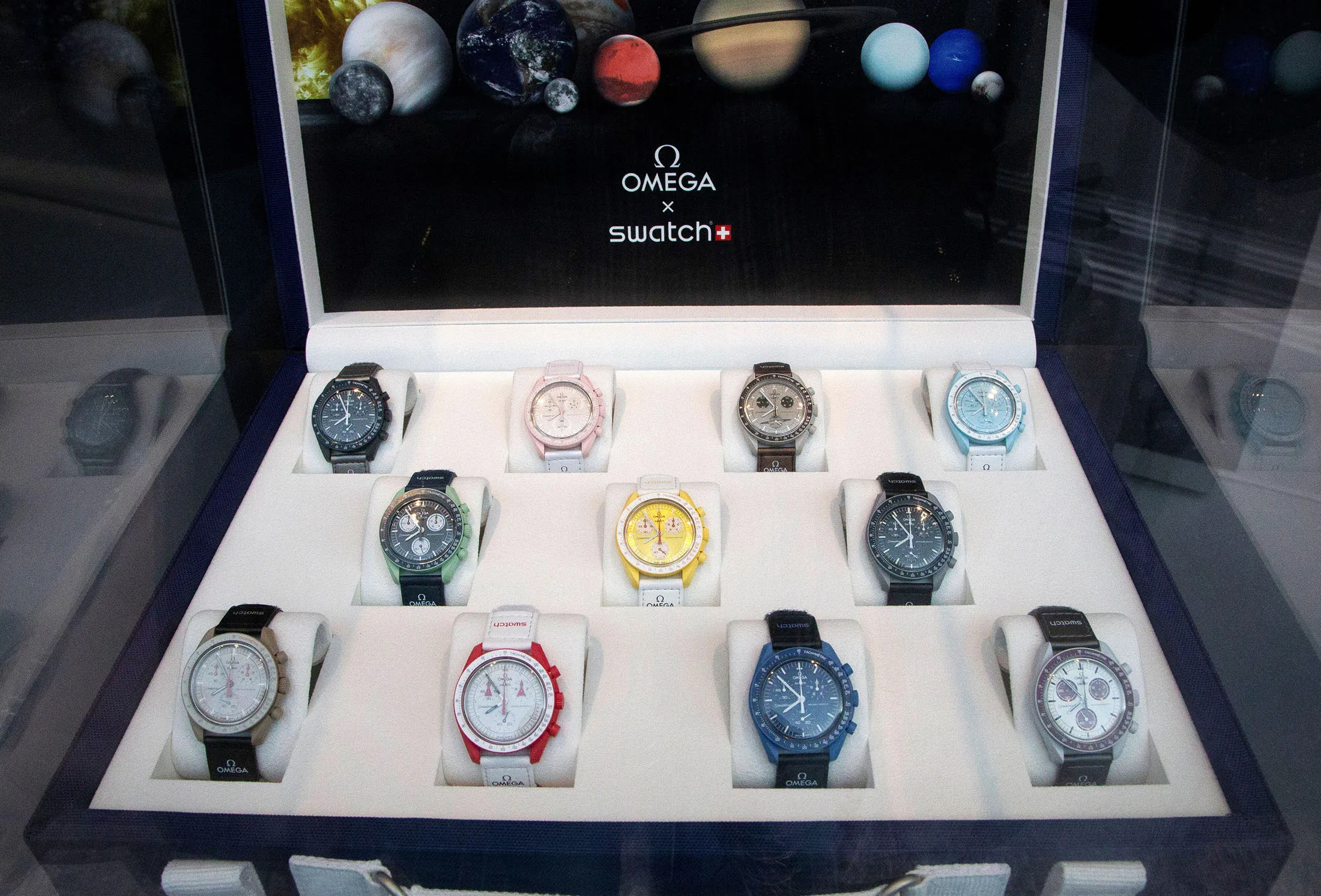 Omega x Swatch MoonSwatch collection displayed inside a luxury retail presentation case featuring multiple tincture bioceramic Speedmaster-inspired watches beneath planetary graphics and collaboration branding