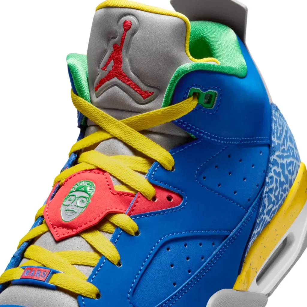 Close-up detail view of a retro basketball shoe highlighting its layered materials and colorful accents. The shoe features a vivid royal blue leather upper with bright yellow laces crossing over a soft gray tongue finished with an embroidered red logo detail. A coral-red lace shroud adds contrast and includes a molded graphic badge accent, while green padded lining frames the collar. Blue elephant-print texturing wraps the heel area and connects to a speckled yellow midsole with visible air cushioning, creating a bold mix of classic basketball heritage and playl street-culture energy
