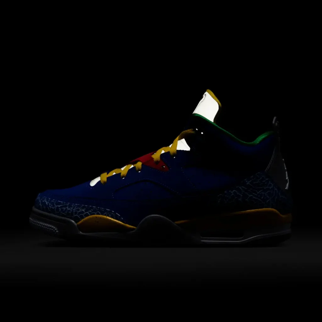 Moody low-light product image of a retro basketball shoe photographed against a deep black background. Most of the silhouette remains in shadow, while selective lighting reveals flashes of vivid royal blue leather, yellow laces, coral-red lace accents, and textured elephant-print overlays around the toe and heel. Reflective details on the tongue and lace area catch the light, creating a dramatic contrast that emphasizes the shoe’s layered construction and bold color palette. The composition gives the sneaker a cinematic, after-dark presentation with a collectible, showcase feel