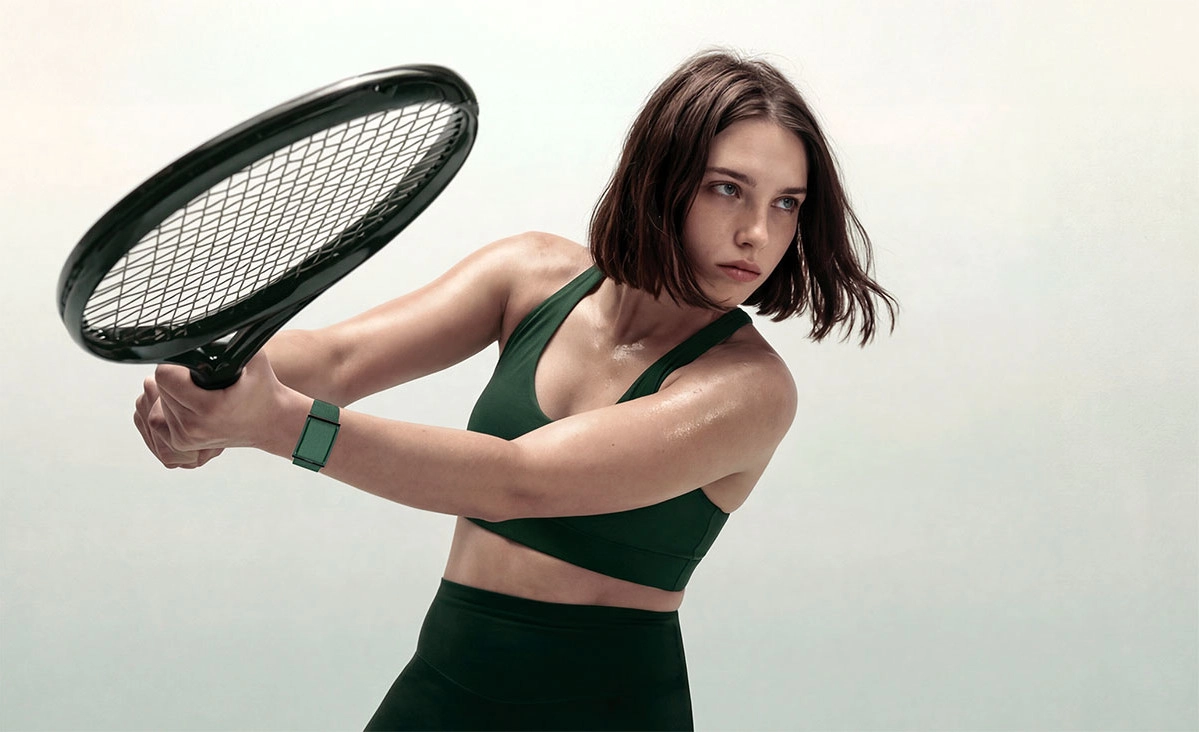 Lifestyle campaign image of a woman mid-swing holding an oversized racket while wearing the green Luna Band on her wrist, highlighting the wearable’s slim, screen-free design and sporty aesthetic