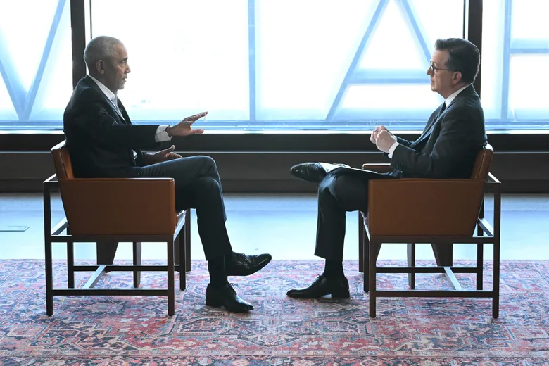 Barack Obama and Stephen Colbert sit across from one another during a televised interview inside the Obama Presidential Center in Chicago, framed by expansive geometric windows and minimalist interiors. Obama gestures mid-conversation while Colbert listens attentively, reinforcing the reflective and conversational tone of the widely discussed 2026 late-night exchange