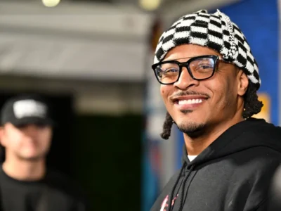 T.I. smiling during a public appearance while wearing black-framed glasses, a black hoodie, and a black-and-white patterned knit cap, with a softly blurred background a guy wearing a cap
