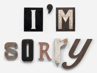 Assorted vintage industrial metal and enamel letters arranged against a pale gray background to spell out the phrase “I’m sorry.” Each character features a different texture, color, and typographic style, ranging from weathered black enamel to rusted steel and painted cream finishes, giving the composition a handmade, salvaged aesthetic with emotional undertones of apology and vulnerability