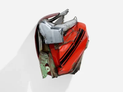 Crushed red and chrome car body fragment with black racing stripes mounted against a pale gray backdrop, showing warped metal, exposed rust, and sculptural industrial textures
