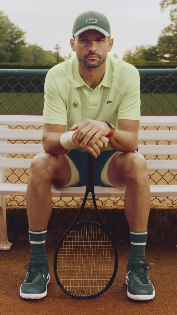 Editorial tennis portrait of a player seated on a white courtside bench with hands resting over a vertically positioned racket, dressed in coordinated Lacoste performance apparel including a pale green polo, green cap, striped socks, and dark court shoes. Set against a chain-link fence and outdoor court backdrop, the image blends heritage tennis styling with a calm, modern campaign aesthetic