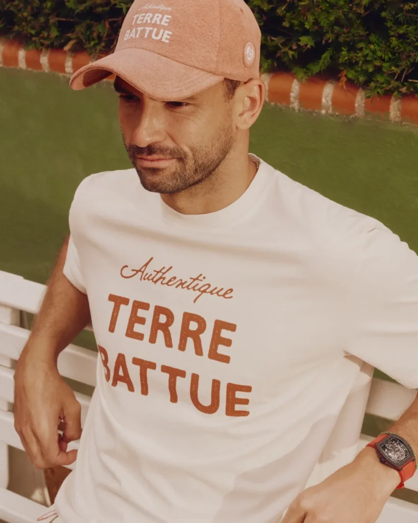 Close-up lifestyle campaign portrait of a tennis-inspired look featuring a soft off-white graphic T-shirt with oversized “Authentique Terre Battue” lettering paired with a matching clay-toned cap embroidered with the same phrase. Seated against a white courtside bench and green backdrop, the styling references French clay-court culture through warm earthy tones and relaxed off-court dressing