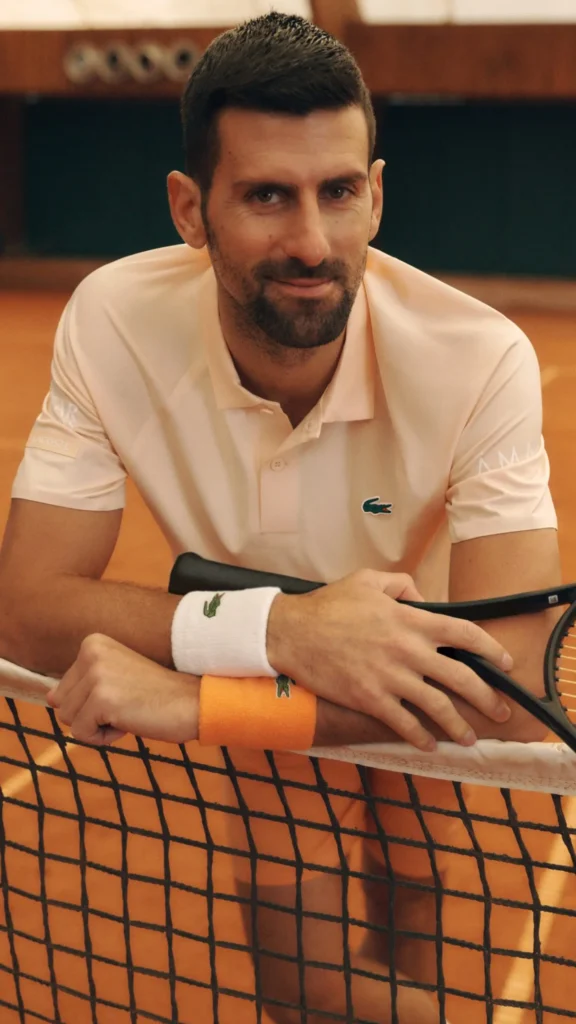 Editorial portrait of a tennis player leaning across the net on a clay court while holding a racket, dressed in a soft blush-pink Lacoste performance polo with coordinated wristbands in white and clay orange. The close framing and warm court lighting create a relaxed yet elevated campaign mood that blends tournament heritage with contemporary sport style