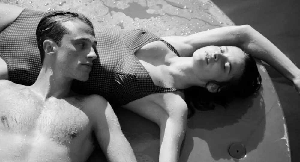 Black-and-white film still showing a man and woman reclining together on a circular floating platform, their skin glistening with water under direct sunlight. The woman rests with eyes closed while the man looks toward her, creating a scene of quiet intimacy, physical stillness, and understated emotional distance against the shimmering surface of the water