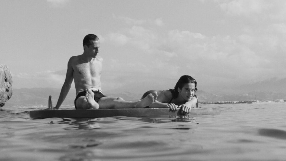 Black-and-white still from The Stranger showing a man and woman resting on a floating platform in calm coastal water beneath a bright sky, with distant mountains and shoreline fading into the background. The scene emphasizes stillness, intimacy, and the film’s sun-soaked yet emotionally restrained atmosphere