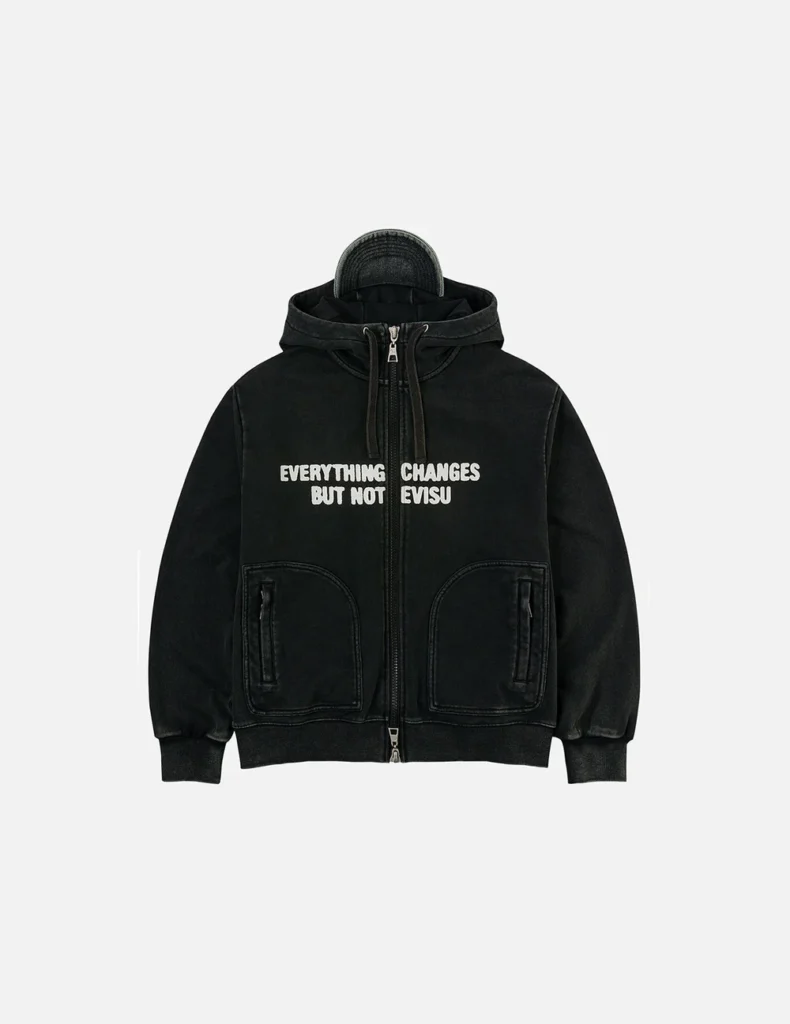 Front-facing product shot of a black Evisu oversized hooded jacket featuring a two-way zip, curved front pockets, ribbed trims, and bold white slogan print reading “Everything Changes But Not EVISU,” with the integrated cap subtly visible within the hood