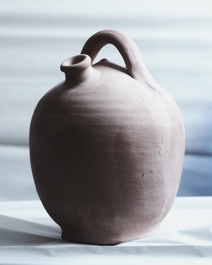 Hand-formed matte ceramic vessel with rounded body, asymmetrical handle, and narrow spout opening, photographed in soft natural light against a muted minimalist backdrop