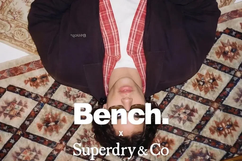 Bench x Superdry & Co. campaign image featuring a model lying upside down on a patterned vintage-style rug while wearing a dark Bench jacket layered over a red plaid shirt and white tee. The flash-lit composition and relaxed styling create a nostalgic early-2000s mood, while large Bench and Superdry & Co. logos overlay the center of the image to emphasize the revived British streetwear identity